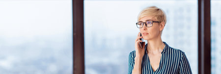 Serious Businesswoman Talking On Cellphone Standing Near Window In Office