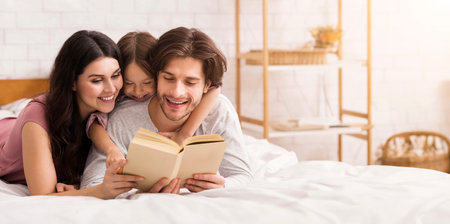 Young parents and their little daughter reading book in bed togetherの写真素材