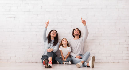Family of three sitting on floor near wall and pointing upwards