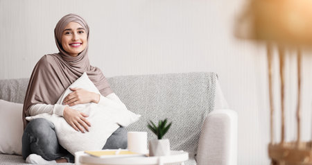 Smiling Muslim Woman In Hijab Sitting On Sofa And Cuddling Pillowの写真素材