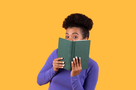 Woman hides playfully behind a green book on a yellow backgroundの写真素材