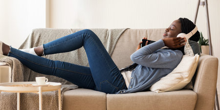 African American girl lying on couch, enjoying music with wireless headphones