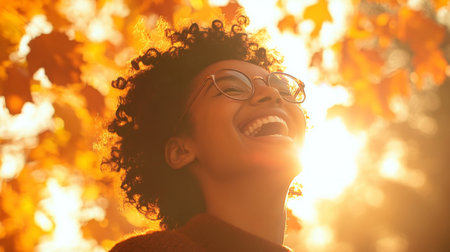 Young woman with curly hair smiles joyfully under vibrant autumn leaves in golden sunlightの素材