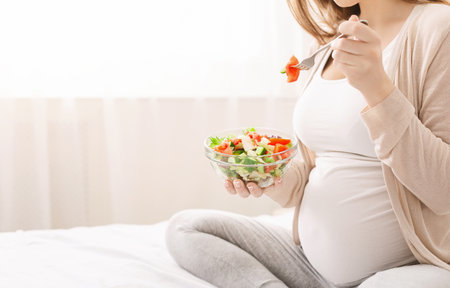 Pregnant woman eating fresh vegetable salad in bedの写真素材
