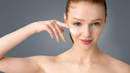Red-Haired Young Woman Applying Cream On Cheeks Over White Backgroundの写真素材