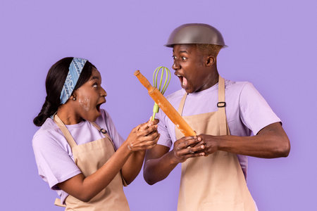 Emotional African American lady and guy fighting with kitchen utensils while cooking on color...