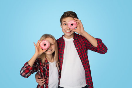 Happy siblings covering eyes with donuts, posing together over blue background