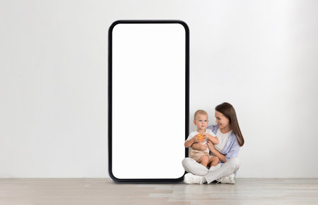 A mother sits with her toddler beside a large blank screen in a bright room