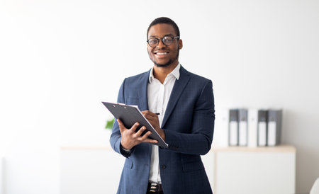 Happy young black male psychologist writing in clipboard, looking at camera and smiling in modern officeの写真素材