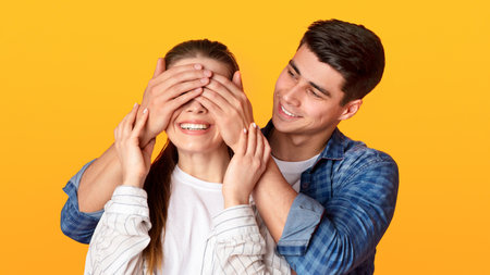 Young guy stands behind lady and closes eyes of smiling woman isolated on yellow backgroundの写真素材