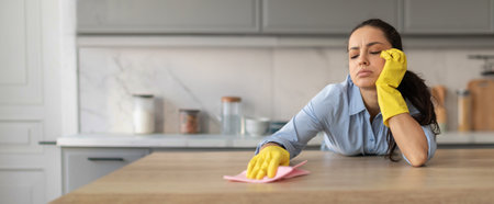 Woman cleaning kitchen countertop while resting her head in hand during taskの写真素材