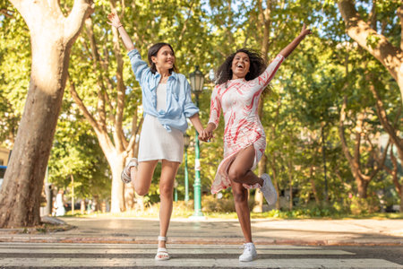 Two friends joyfully leaping while holding hands in a park during daylightの写真素材