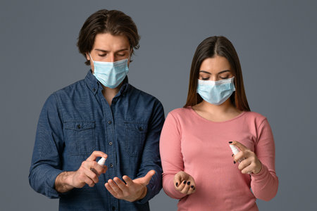 Man and woman in medical masks applying disinfectant spray on handsの写真素材