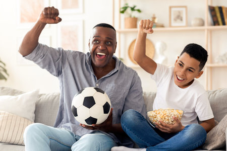 Joyful african grandfather and hid grandson cheering while watching football on tvの写真素材