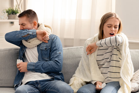Young man and woman coughing into their elbows at homeの写真素材