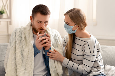 Caring wife offering warm tea to her sick husband at homeの写真素材
