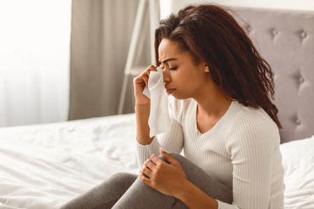 Black woman crying, holding paper tissue sitting in bedの写真素材