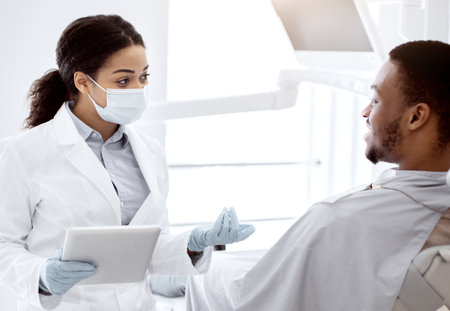 Black Dentist Lady With Digital Tablet In Hands Consulting Male Patient In Clinicの写真素材