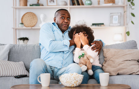 Scary movie night. Senior African American man with his little granddaughter closing face in horror at homeの写真素材
