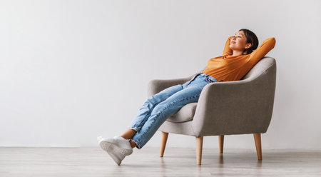 Young Asian woman sitting in comfy armchair, being lazy, feeling peaceful against white wall, panorama with copy spaceの写真素材