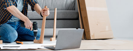 Millennial guy fixing furniture, repairing wooden table uses electric drill, sits on floor at homeの写真素材