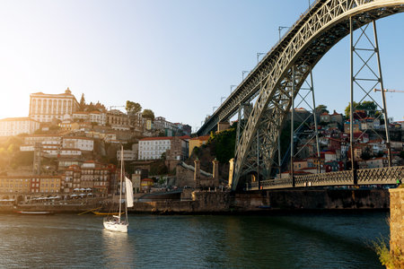 Sailboat gliding under the iconic bridge of Porto at sunset with colorful houses
