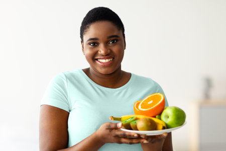 Smiling overweight African American woman holding plate of fresh fruits, keeping healthy diet for weight loss at homeの写真素材