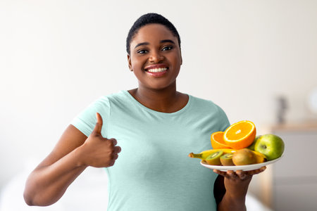 Happy plus size black woman holding fruits and showing thumb up, recommending healthy diet for weight loss at homeの写真素材