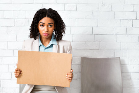 Young black lady with blank sign waiting for employment interview at office lobby, space for designの写真素材