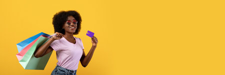 Seasonal sales concept. Happy black woman with colorful shopping bags and unlimited credit card over yellow backgroundの写真素材