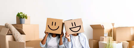 Moving and real estate concept. Black couple with cardboard boxes with smiley faces on head, sitting on floorの写真素材