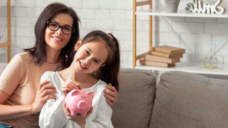 Happy young mother and her teen daughter holding piggybank, sitting on sofa at home, copy spaceの写真素材