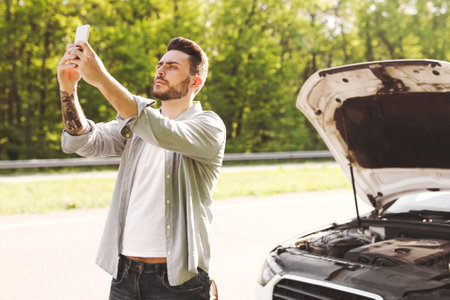 Young guy standing near broken car after accident, looking for mobile network signal, trying to call roadside assistanceの写真素材