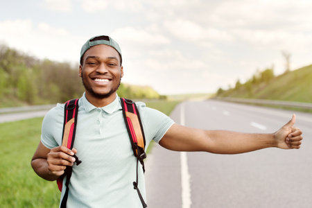 Smiling black guy with backpack flagging down car, catching free ride, hitchhiking on highway, free...