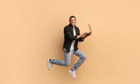 Young man joyfully using laptop while jumping in front of a solid background