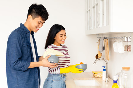 Asian guy helping his cheerful girlfriend washing dishes at kitchenの写真素材
