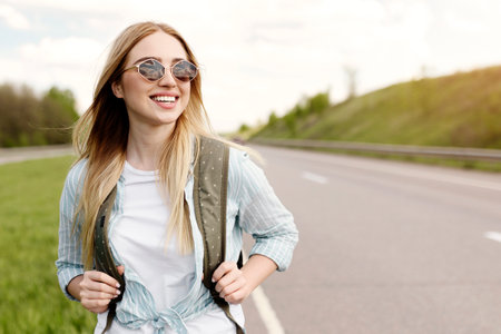 Cheery young lady in casual summer clothes walking along road in countryside, hitchhiking for ride outdoorsの写真素材