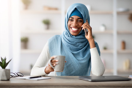 Cheerful black muslim woman talking on cellphone and drinking coffee in officeの写真素材