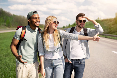 Cheerful diverse friends trying to get free ride, hitchhiking on road, showing thumb up outdoors