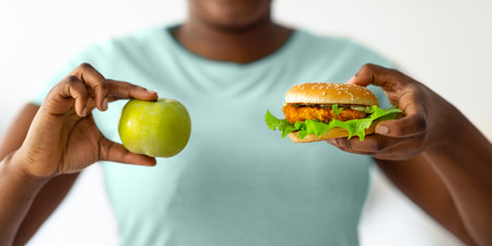 Unrecognizable overweight black woman choosing between burger and apple, closeup. Nutrition for weight lossの写真素材