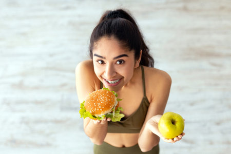 Above view of happy young Indian woman holding apple and hamburger, standing on scales, choosing her dietの写真素材