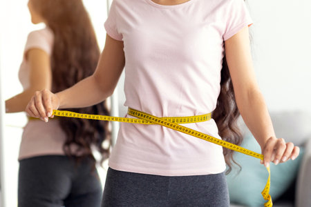 Unrecognizable Indian lady measuring her waist with tape in front of mirror at home, closeupの写真素材