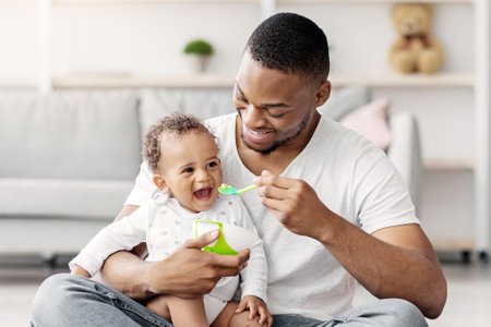 Happy Young Black Dad Spoon Feeding His Adorable Baby Son At Homeの写真素材