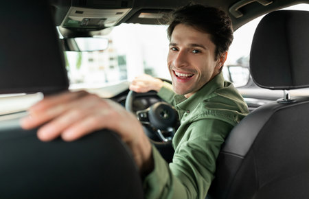 Cheerful young man driving new automobile after purchase, sitting in drivers seat of car at...