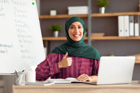 Islamic teacher in headscarf sitting at desk showing thumbs upの写真素材