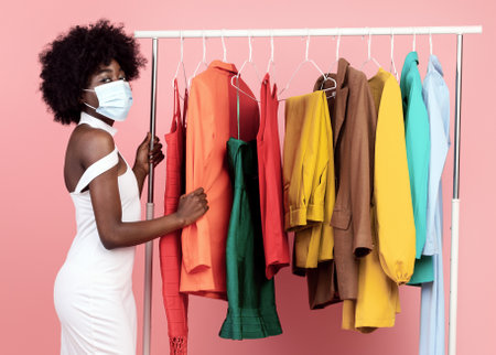 Black Lady Wearing Face Mask Choosing Clothes Over Pink Backgroundの写真素材