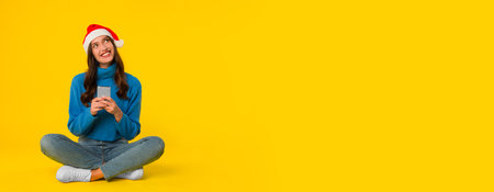 Cheerful lady in Santa hat texting on cellphone, yellow backgroundの写真素材