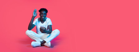 A young man sits on a pink background and waves with a phone in his handの写真素材