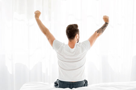 Relaxed young man waking up in bed and stretching his arms, facing the window, back view, free spaceの写真素材