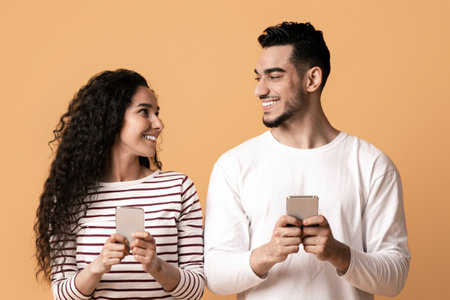 Cheerful Arab Man And Woman Holding Smartphones And Looking At Each Other